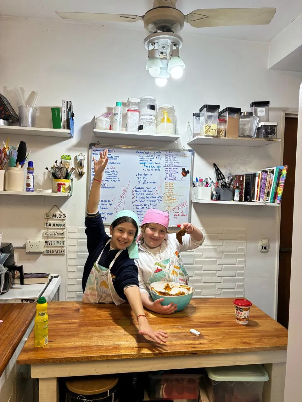 Dos chicas cocinando en la cocina de Mariana, con el pizarrón de producción atrás