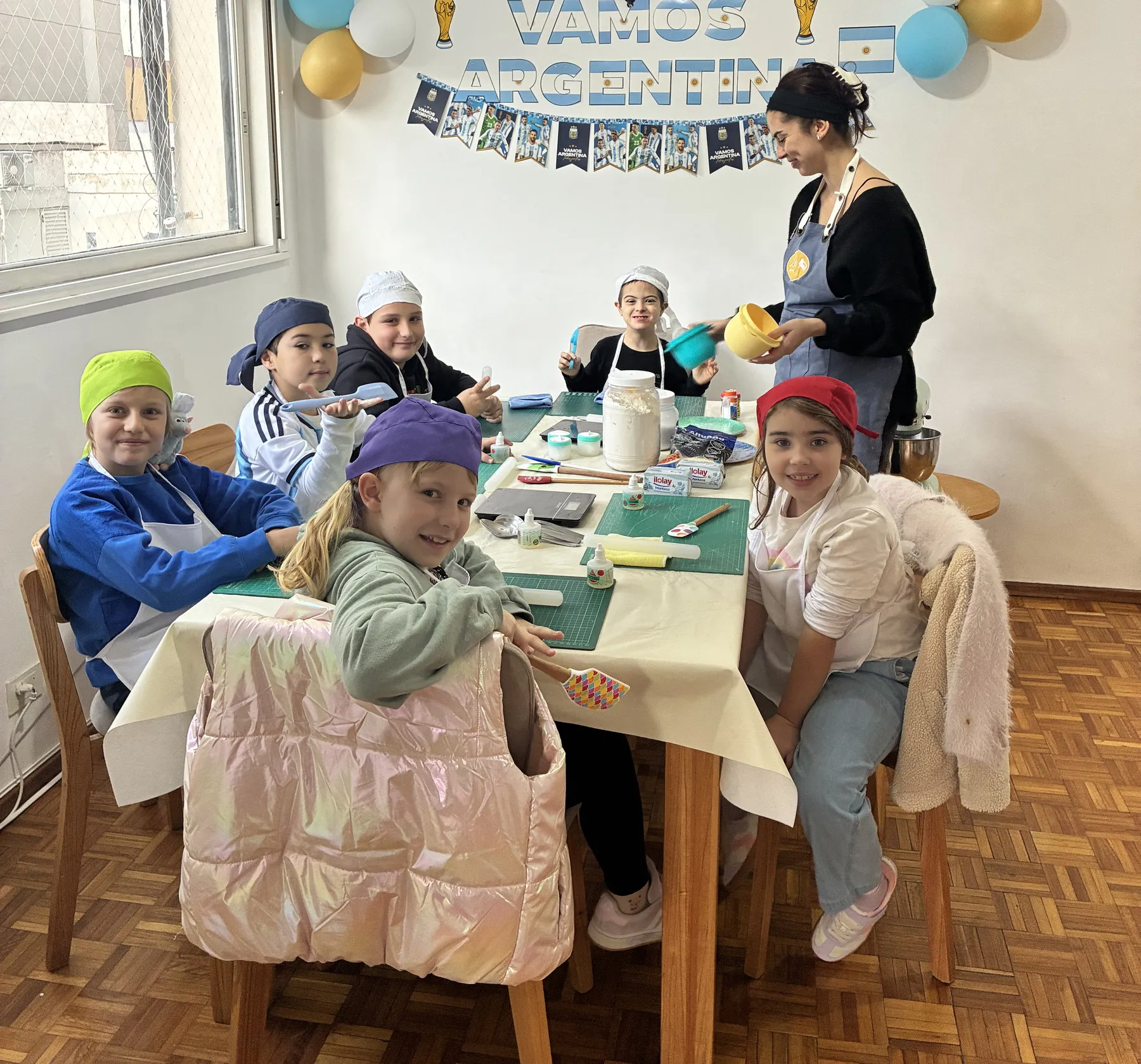 Seis chicos con delantal y gorro alrededor de la mesa de trabajo, con alguien del equipo de Lottie's sirviendo los ingredientes